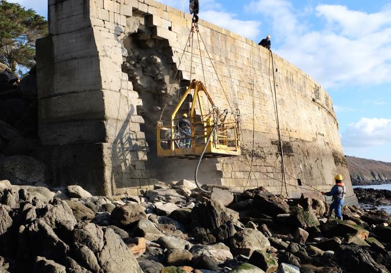 Vue des travaux de consolidation de 2015 : du béton est projeté sur la maçonnerie de la brèche de la batterie basse par des techniciens travaillant avec une nacelle suspendue